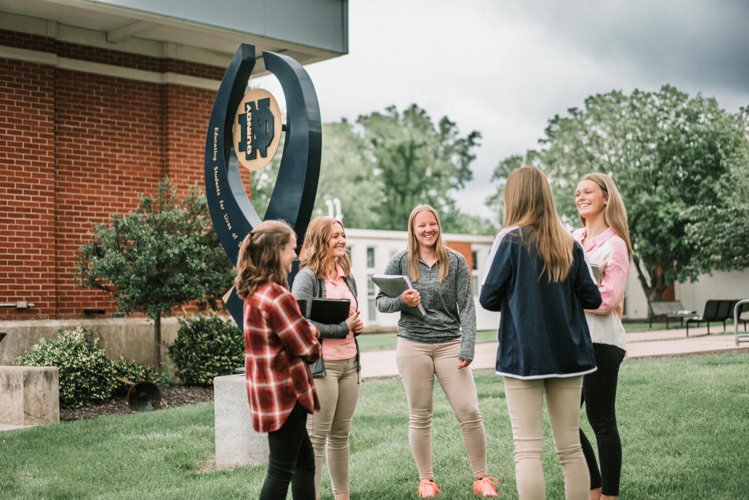 Group of QND students smiling and chatting near a campus sculpture at Quincy Notre Dame High School