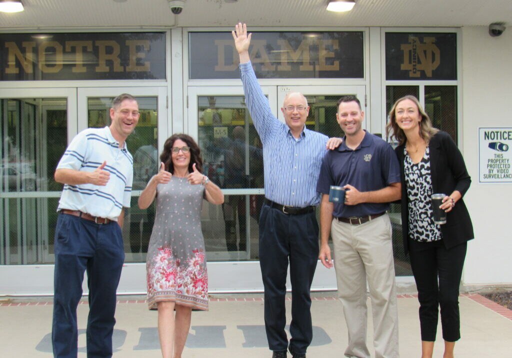 QND Faculty and Staff - Smiling QND faculty and staff posing outside Quincy Notre Dame High School entrance, showing Raider Pride and teamwork