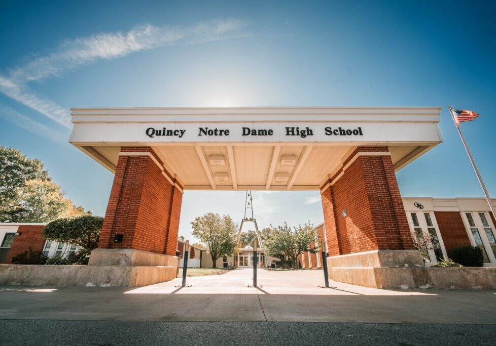 QND Overview / FAQ - Front entrance portico of Quincy Notre Dame High School with clear blue sky and American flag